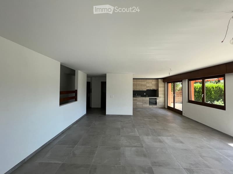 Empty room with grey tiled floor, white walls, and a small kitchen area visible in the background. No furniture or personal items are present.