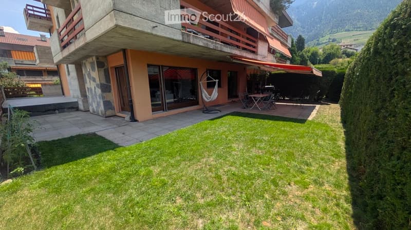 Well-maintained garden with green grass, patio area with outdoor furniture, and a modern house with balconies and large windows in the background.