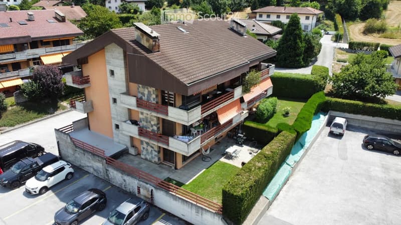 A modern multi-story house with a tiled roof, balconies, and a well-maintained garden. The house features a mix of stone and wood exterior finishes, with several parked cars in the driveway.
