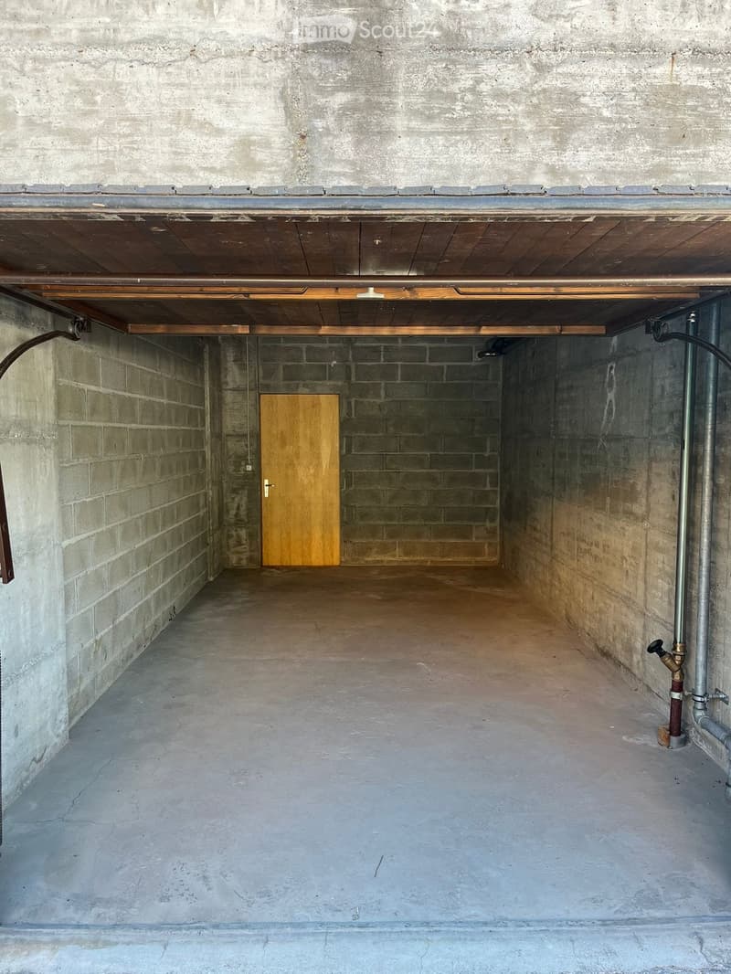 Empty underground garage with concrete walls and floor, wooden door, exposed pipes, and a wooden ceiling with lighting.