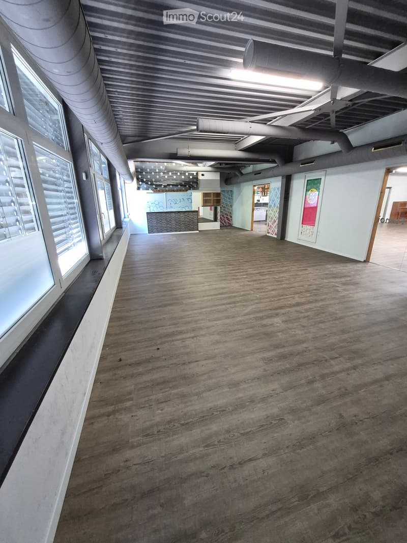 Empty room with wooden flooring, large windows, exposed ceiling pipes, and a small reception area visible in the background.