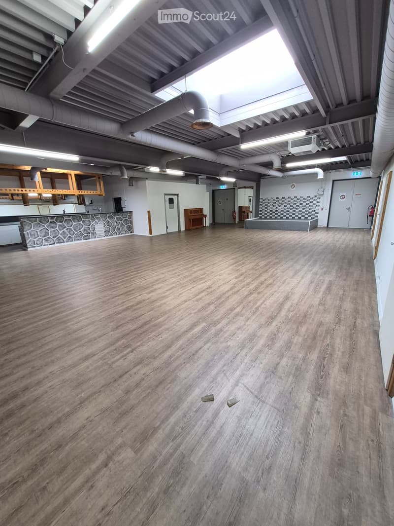 Empty room with wooden flooring, white walls, and exposed industrial-style ceiling with pipes and lighting. Features include a hexagonal-patterned counter, a small seating area, and a doorway leading to another room.