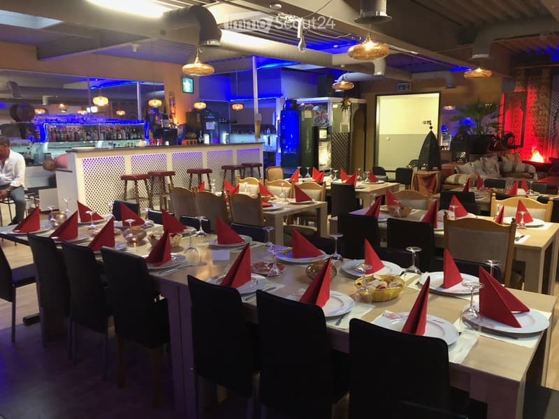 Restaurant interior with multiple tables set for dining, each with plates, wine glasses, and red napkins. Stools are arranged around a bar area, and a refrigerator is visible in the background.