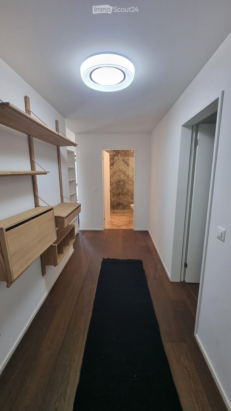 Empty hallway with wooden floor, white walls, ceiling light, wooden shelves, black rug, open door to bathroom