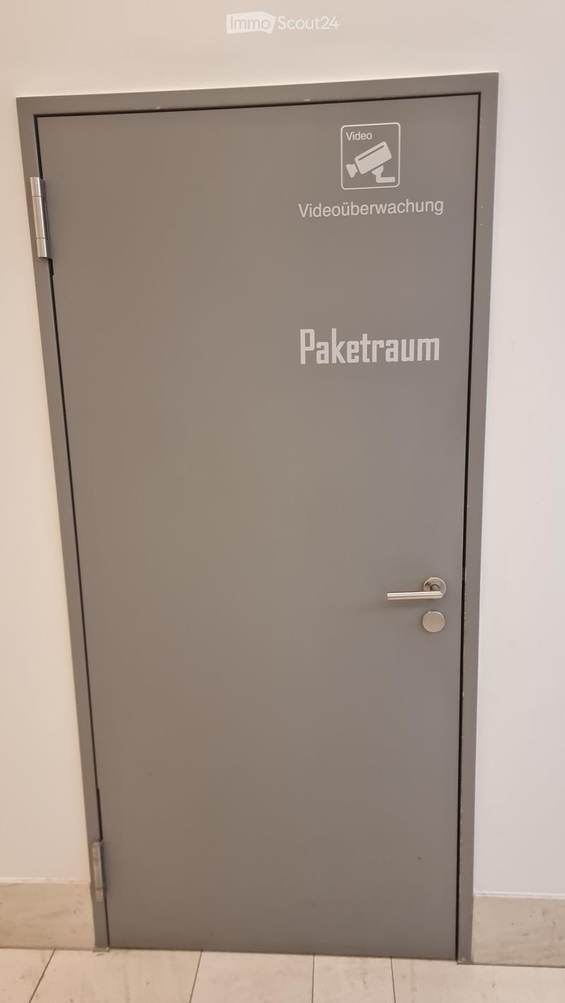 Gray door with silver handle, labeled 'Paketraum' and 'Videoüberwachung'.