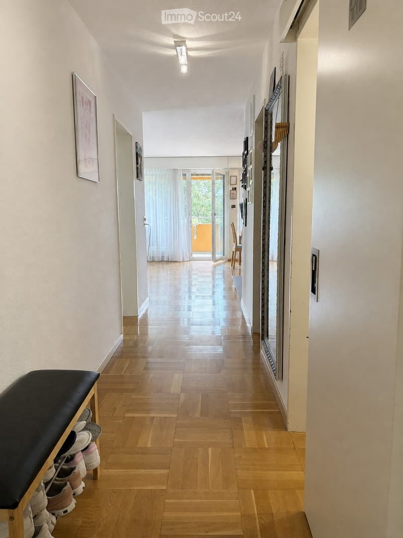 A well-lit hallway with wooden flooring, white walls, and several framed pictures. A bench with shoes is placed on the left side, and a large mirror is mounted on the right wall. The hallway leads to an open doorway with sheer curtains, revealing a glimps