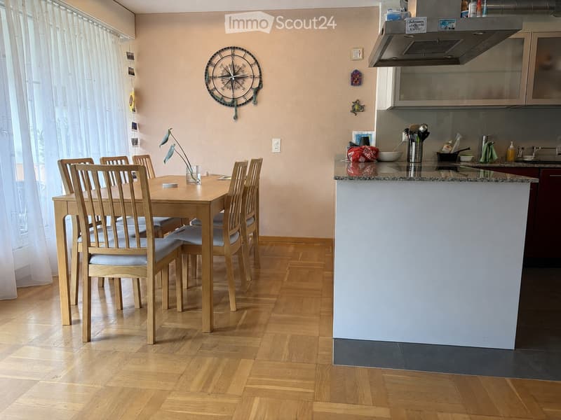 A modern, well-lit dining and kitchen area with a wooden dining table surrounded by chairs, a kitchen island with a sink and faucet, and a decorative clock on the wall.