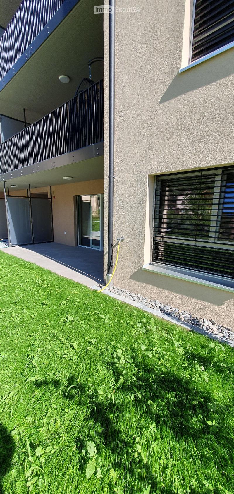 Garden area with green grass, small plants, and a modern building with a balcony and windows.