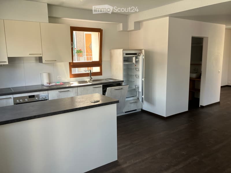 Modern kitchen with white cabinets, stainless steel appliances, and a central island. Features an open refrigerator, sink with faucet, and a window overlooking a balcony.