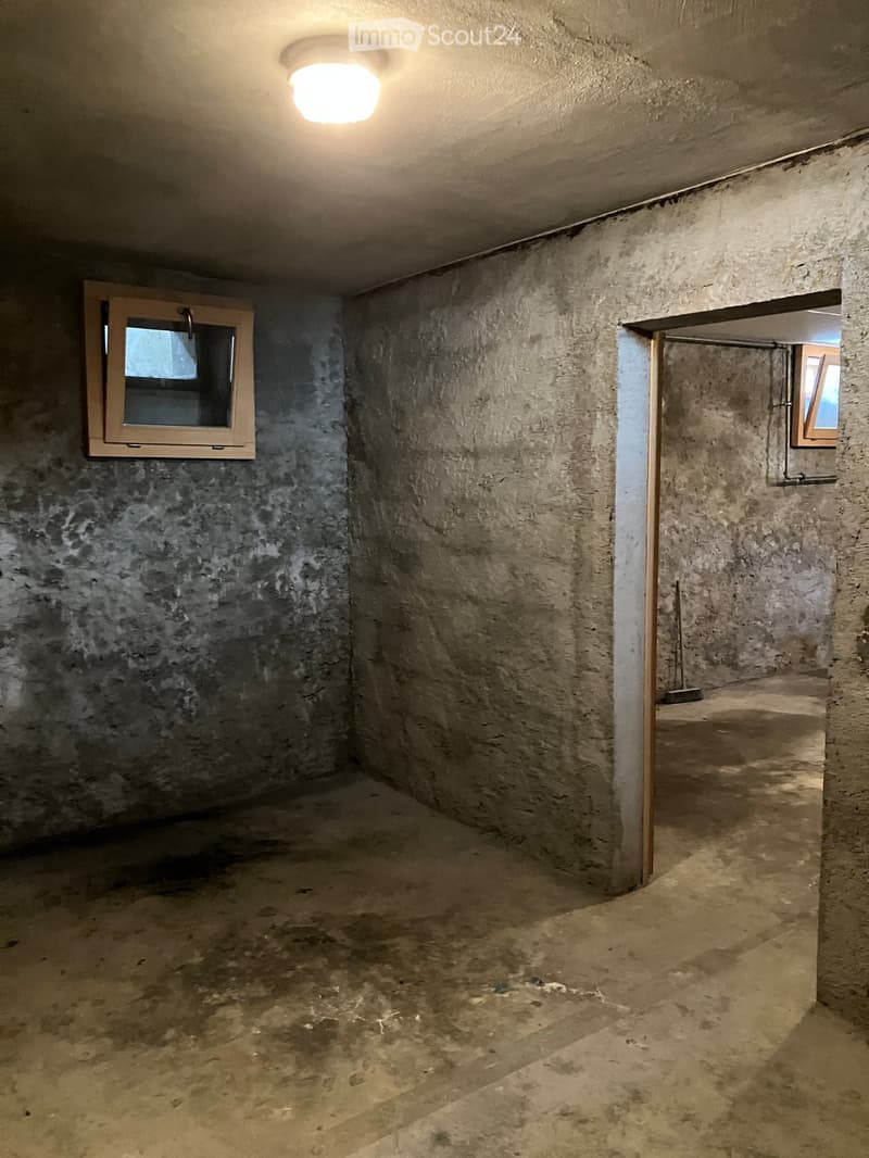 Empty basement room with concrete walls, a single window, and a small doorway leading to another area. The floor is unfinished concrete.