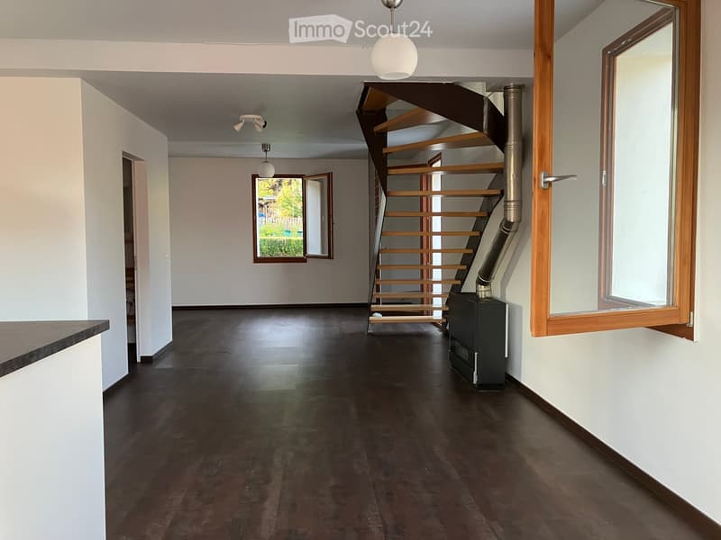 Empty hallway with dark wooden flooring, white walls, and a modern staircase leading upwards. Features include a window with a view of greenery, a ceiling light fixture, and a small black appliance near the staircase.
