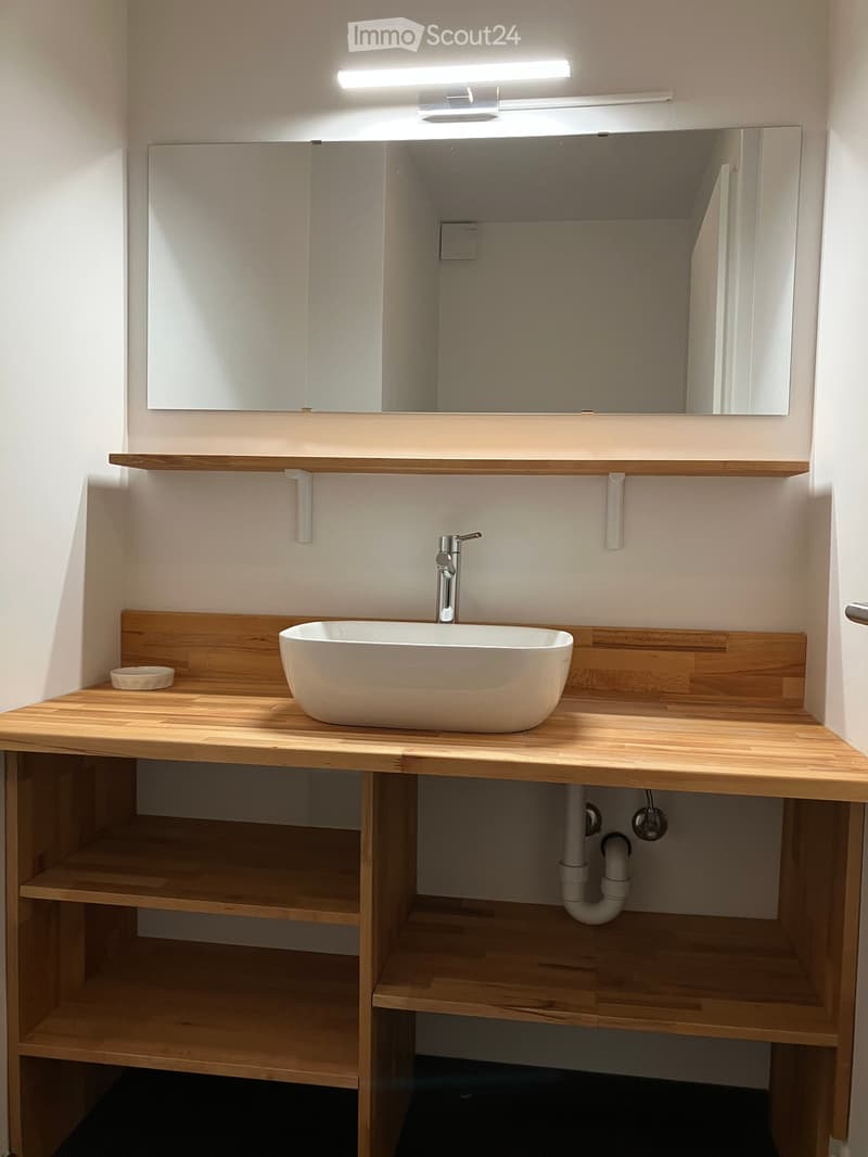 Modern bathroom with a wooden vanity, white sink, sleek faucet, and large mirror above. The vanity has built-in wooden shelves below.