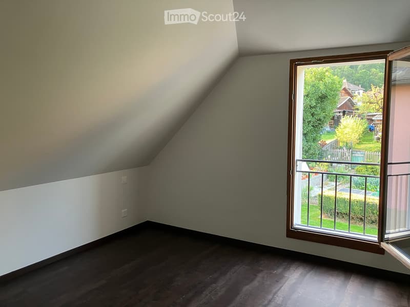 Empty attic room with wooden flooring, slanted ceilings, and a large window offering a view of the garden and surrounding area.