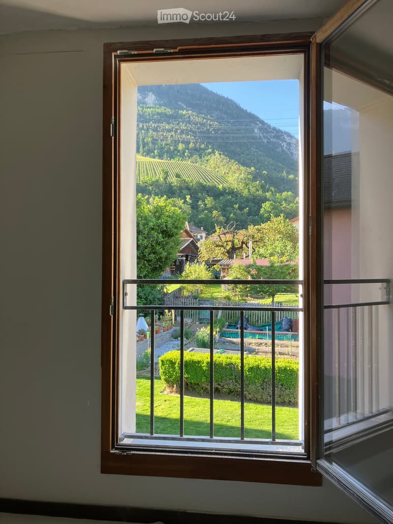 Open window with metal railing overlooking a scenic valley, green fields, and distant mountains.