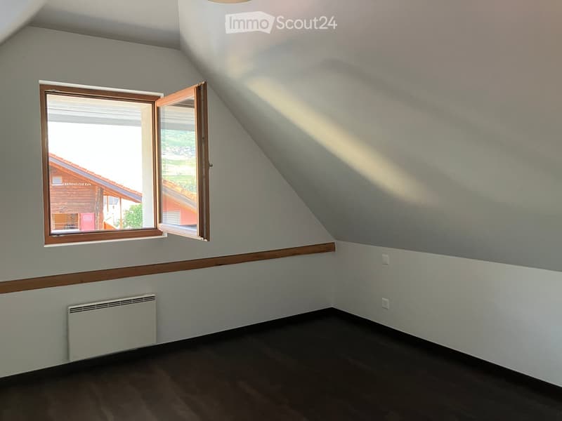 Empty attic room with wooden floor, light grey walls, large window with wooden frame, radiator, and visible electrical outlets.
