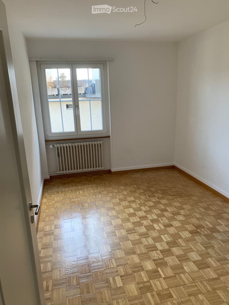 Empty room with wooden parquet flooring, white walls, single window, radiator, and a door.