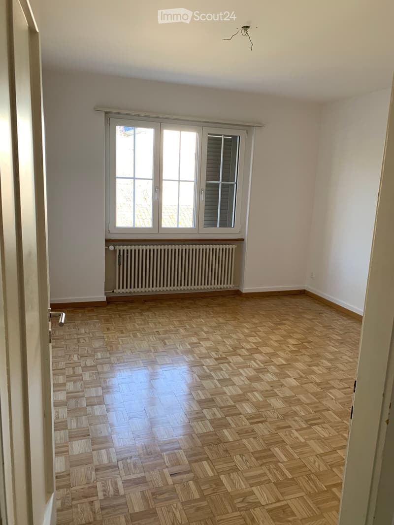 an empty room with wooden parquet floor, large windows, a radiator, and an open door leading to another room