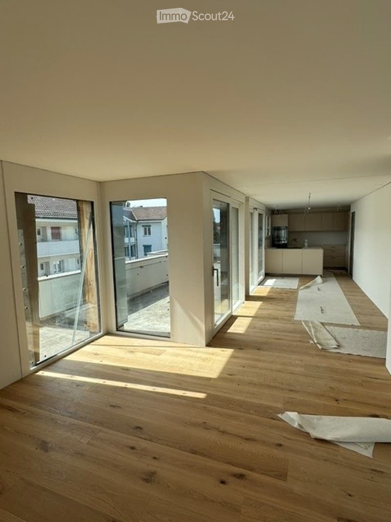 Bright, unfurnished room with wooden flooring, large glass doors leading to a balcony, and a view of neighboring buildings.