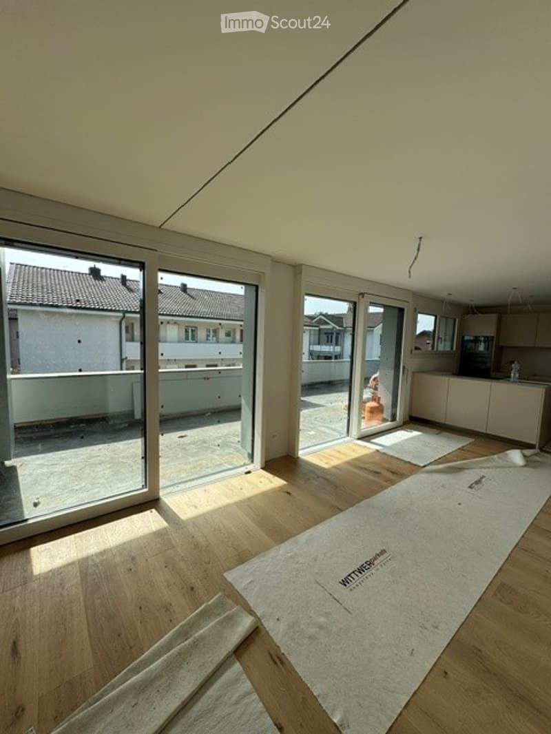 Empty room with wooden flooring, large glass sliding doors leading to a balcony, and a small kitchen area visible in the background.