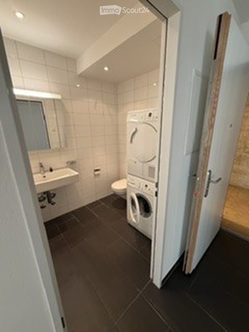 Modern bathroom with a dark tiled floor, white tiled walls, a sink with a mirror, a toilet, and a stacked washing machine/dryer unit.