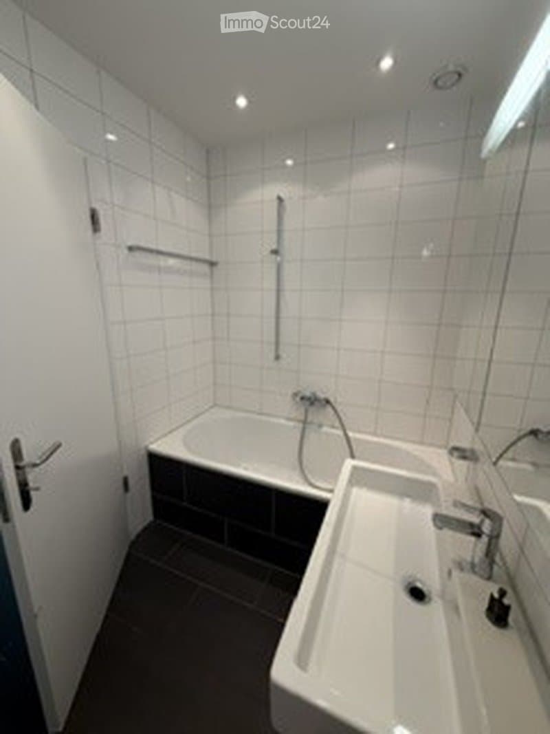Modern bathroom with white tiled walls, a bathtub, a sink, and a mirror. Features include a glass shower enclosure and a door.