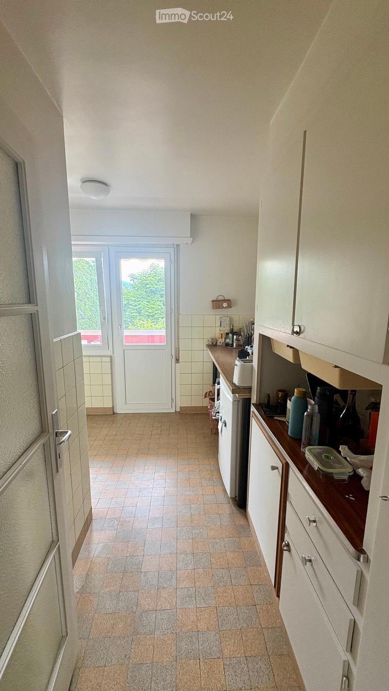 A small kitchen with white cabinets, a wooden countertop, and various appliances including a refrigerator, oven, and toaster. The floor is tiled, and there is a door leading outside.