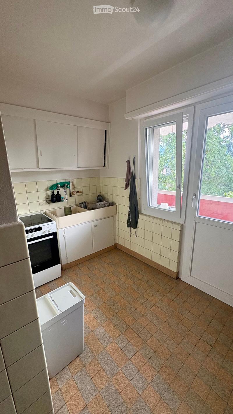 A small kitchen with white cabinets, a tiled backsplash, and a tiled floor. Features include a refrigerator, oven, sink, dishwasher, and a window with a view of greenery.