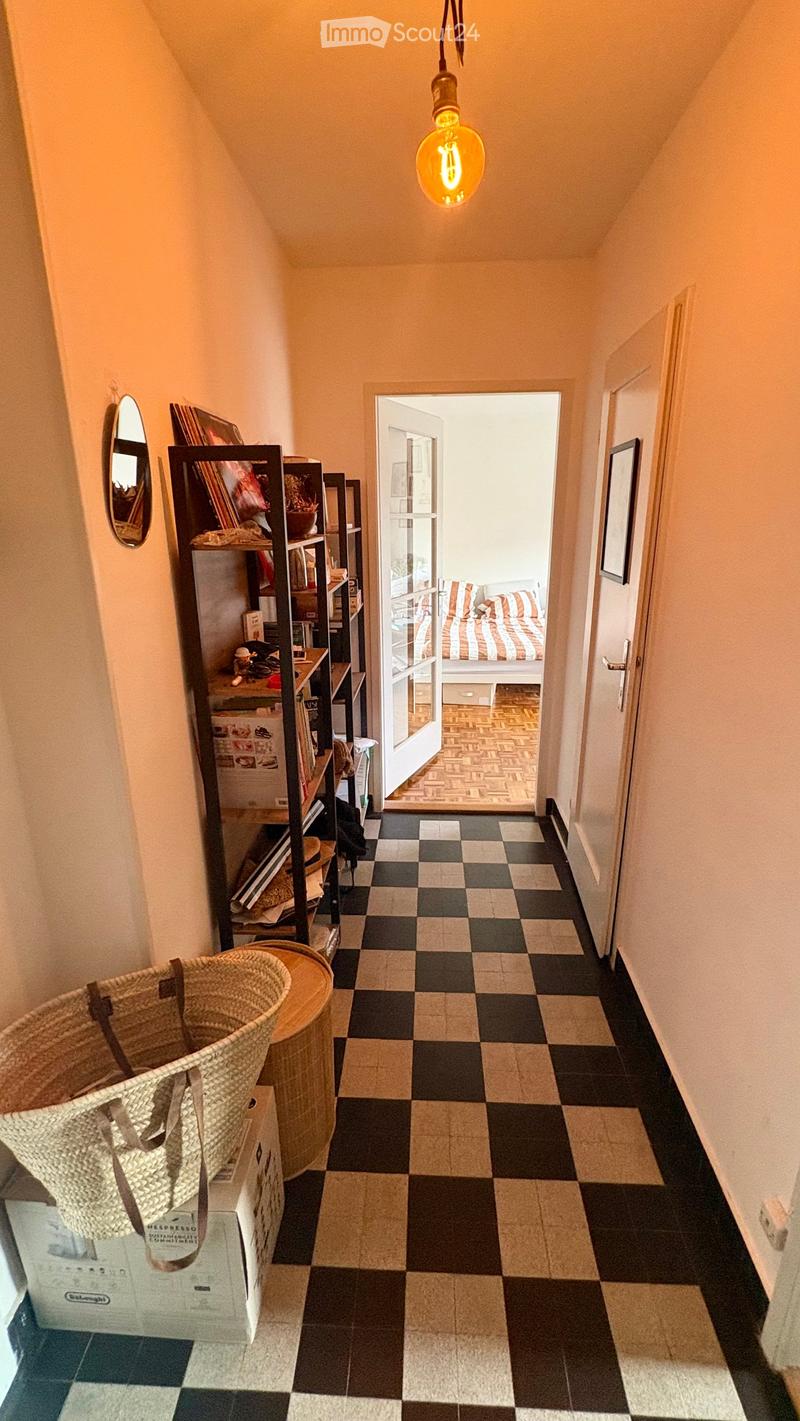 Hallway with checkered black and beige tiles, a hanging light fixture, a shelf with books and items, a mirror, and an open door leading to a bedroom with a bed and striped bedding.