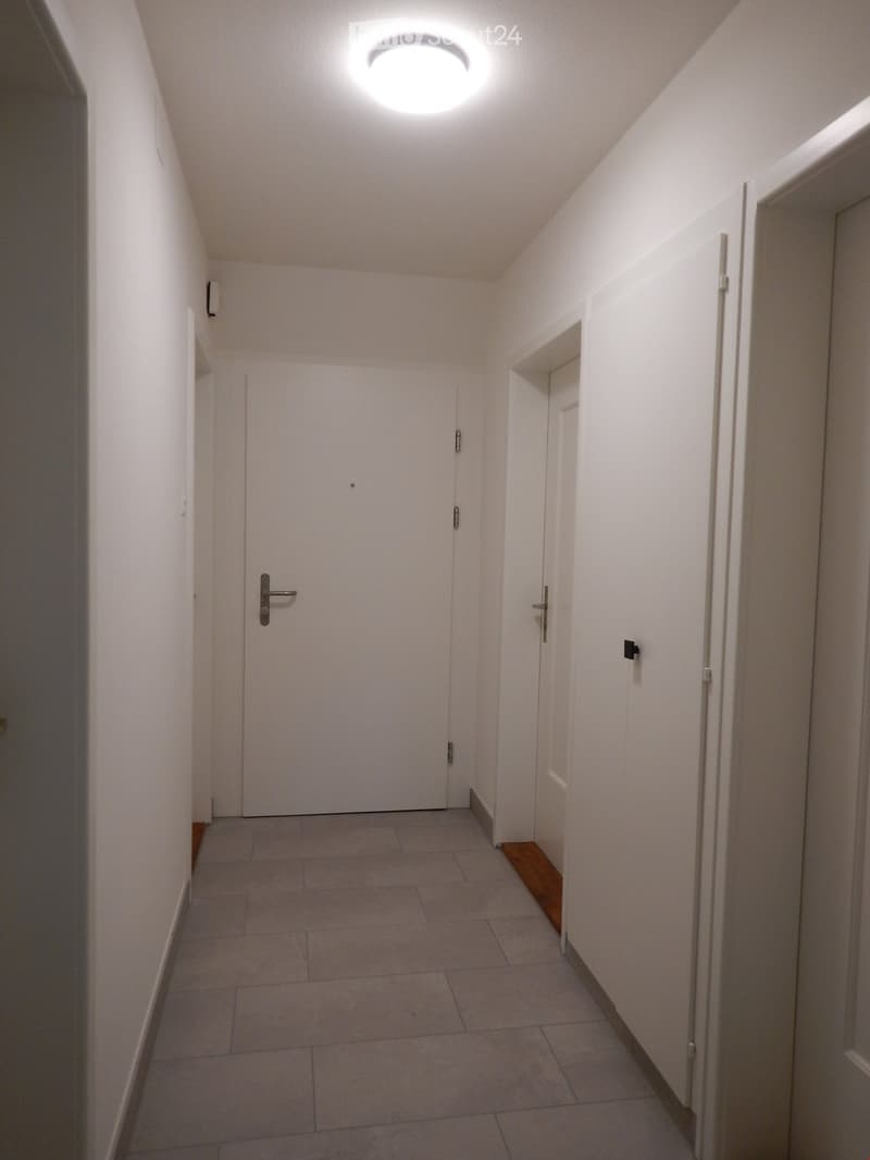 A narrow, well-lit hallway with white walls, tiled floor, and two doors at the end. The ceiling features a round recessed light fixture.