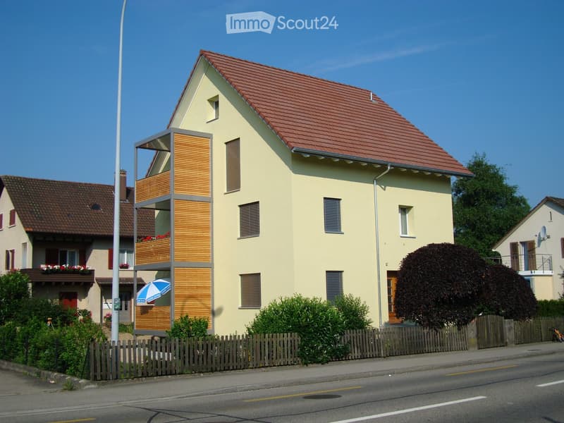 Two-story house with a red-tiled roof, light yellow exterior, and a wooden balcony with a privacy screen. The house features multiple windows, a small garden with trimmed bushes, and a wooden fence along the sidewalk.