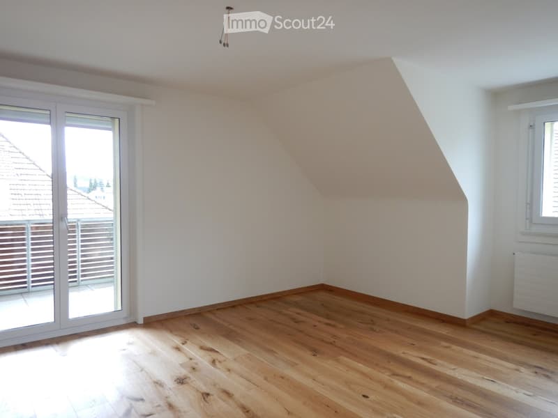 Empty room with wooden flooring, white walls, and large windows, including a sliding glass door leading to a balcony.