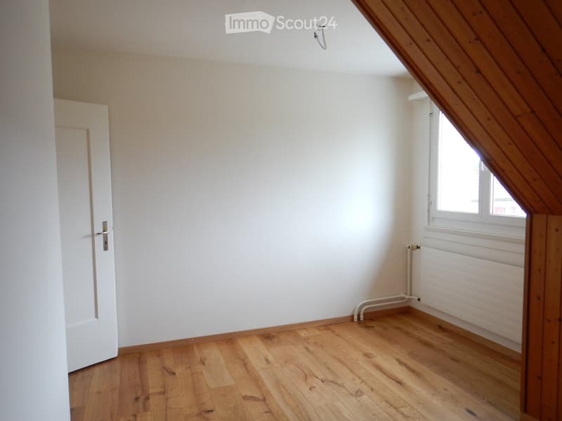 Empty attic room with wooden flooring, white walls, a single window, and a radiator. No furniture or personal items are present.