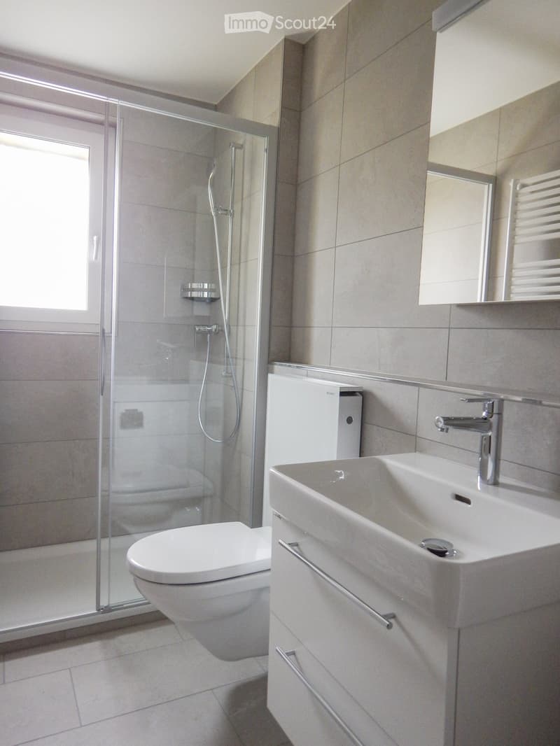 Modern bathroom with a toilet, sink, mirror, and glass-enclosed shower. Light gray tiles and white fixtures.