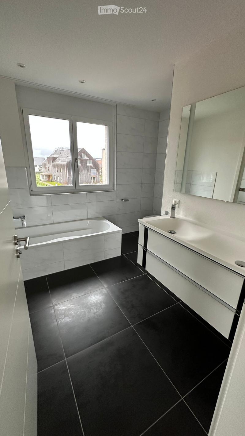 Modern bathroom with black tiled floor, white bathtub, white sink, and large mirror. Large window offers a view of the neighborhood.