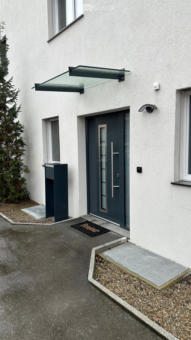 Modern building entrance with a grey door, glass canopy, and a small doormat reading "Bonjour".