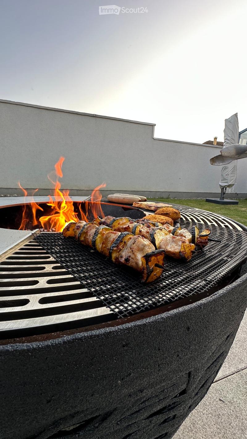 A barbecue grill with skewers of meat and vegetables cooking over an open flame, placed on a concrete surface with a building in the background.