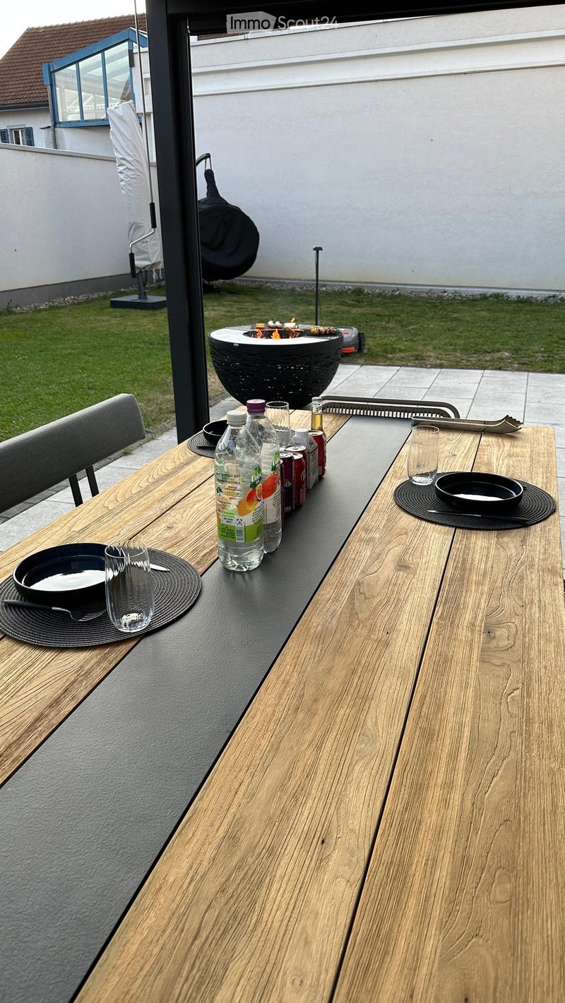 A wooden outdoor table with a black surface, set with bowls, cups, and bottles, accompanied by a chair and a fire pit in the background.