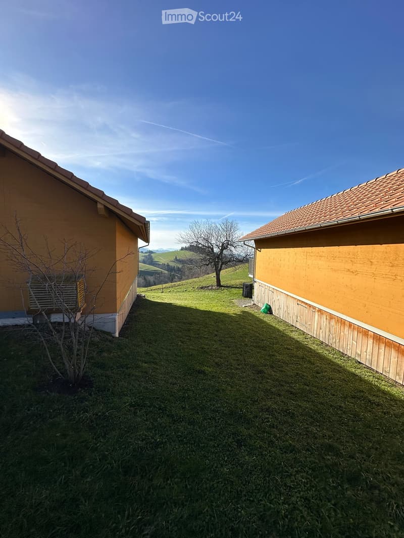 A grassy yard between two yellow buildings with tiled roofs, surrounded by a rural landscape with hills and a clear blue sky.