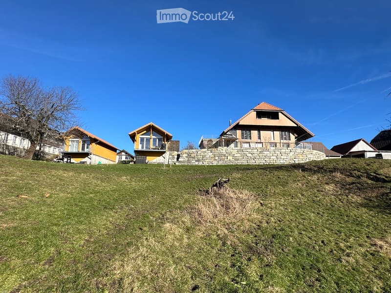 Residential houses with pitched roofs and balconies, surrounded by a grassy slope and stone wall.