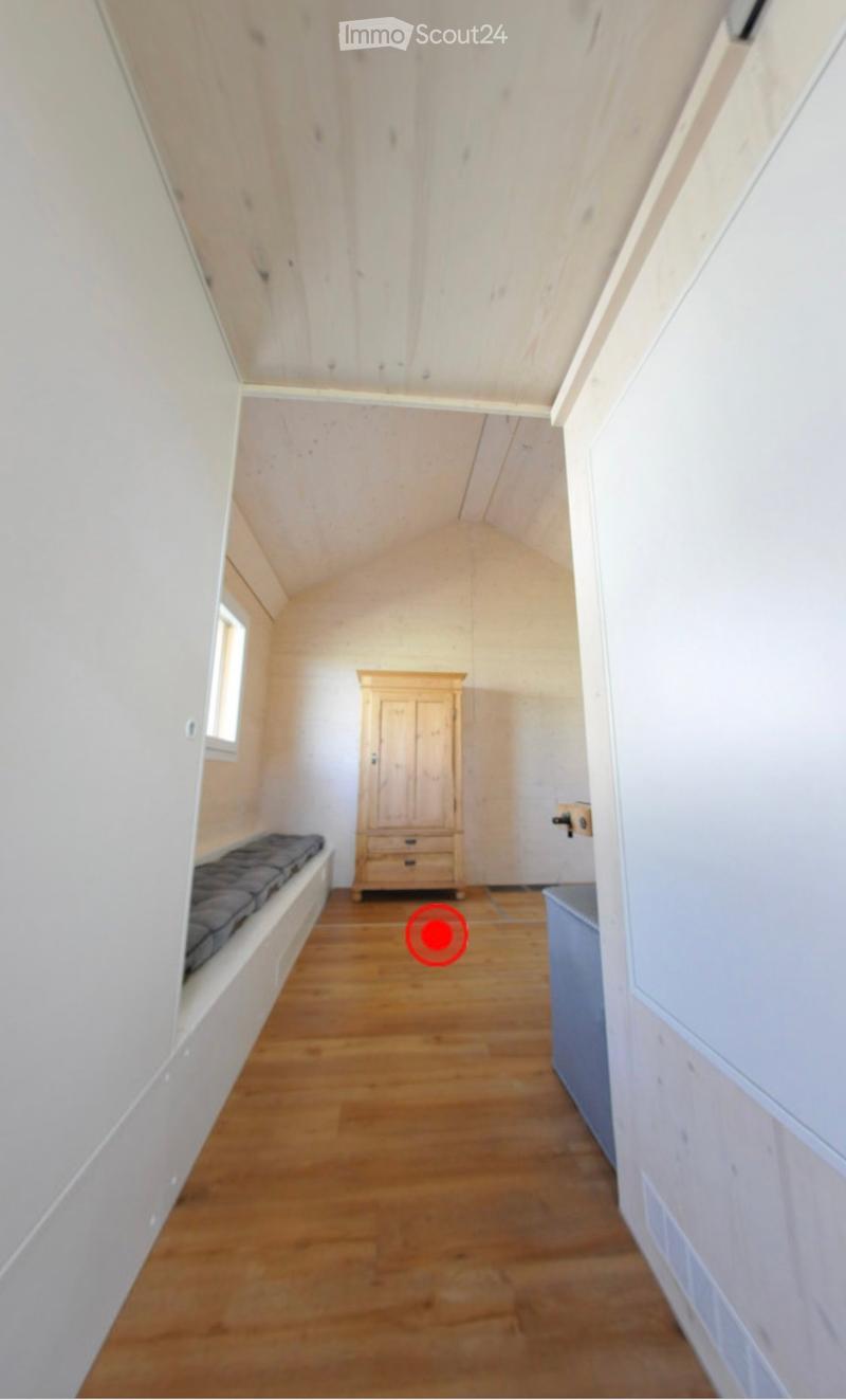 A small, furnished room with wooden flooring, white walls, and a slanted ceiling. Features include a wooden wardrobe and a built-in bench along one wall.