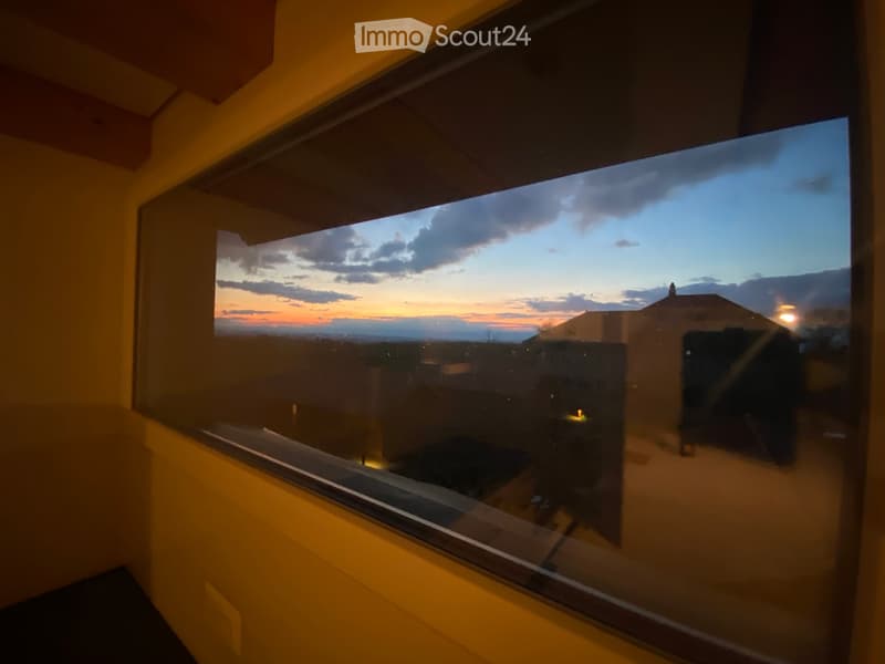 View from a large window showing a sunset over a landscape with a house and outdoor lighting.