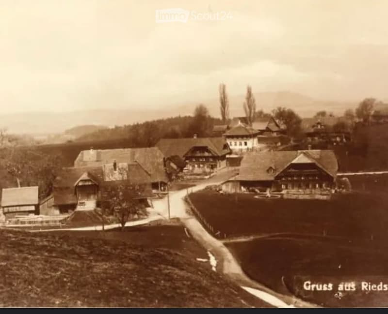 A sepia-toned photograph of a rural village with traditional wooden houses, pitched roofs, and a winding road through a green landscape.