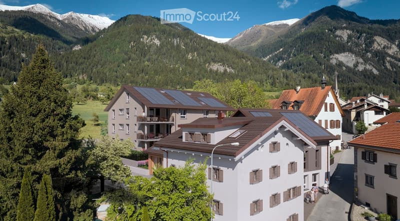 Aerial view of a residential area with multiple houses, solar panels on roofs, surrounded by lush greenery and mountains in the background.