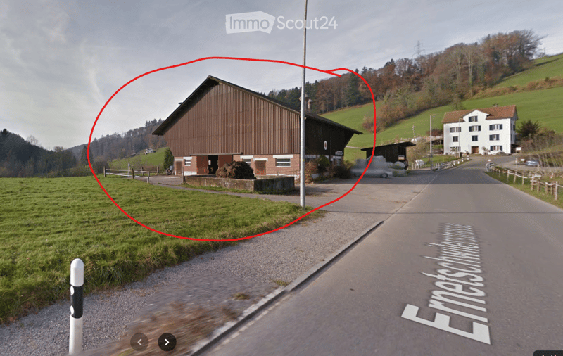 A rural building with a brown exterior and a flat roof, situated on a grassy area near a road, surrounded by green hills and trees.