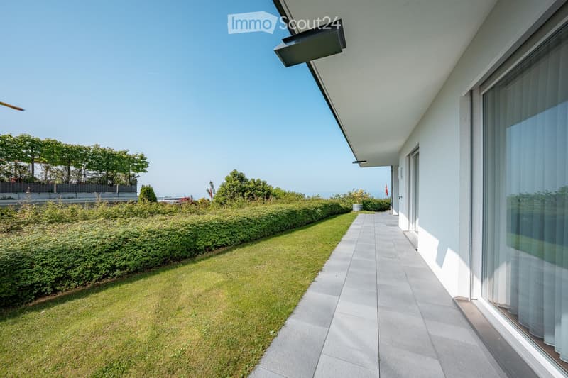 A modern outdoor area with a paved pathway leading to a grassy lawn, surrounded by neatly trimmed hedges and trees, with a view of the sea in the background.