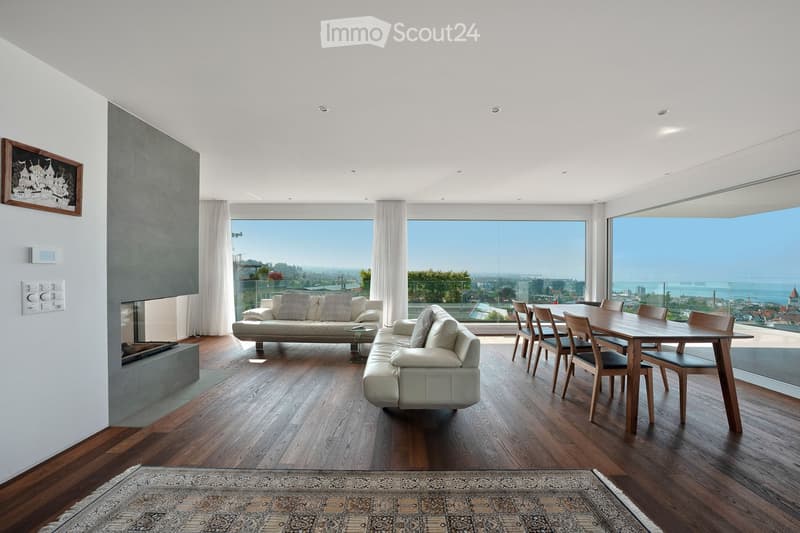 Modern living room with large windows offering a scenic view, featuring a sofa, chairs, a dining table, and a fireplace.