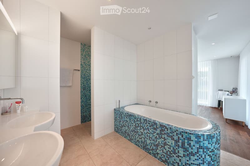 Modern bathroom with a large bathtub featuring blue mosaic tiles, two sinks, and a towel hanging on a rack. The space is bright with white tiles and recessed lighting.