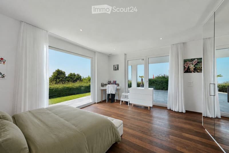 Bright bedroom with wooden flooring, large windows, and a view of greenery. Features a bed, nightstand, chair, desk, and mirror.