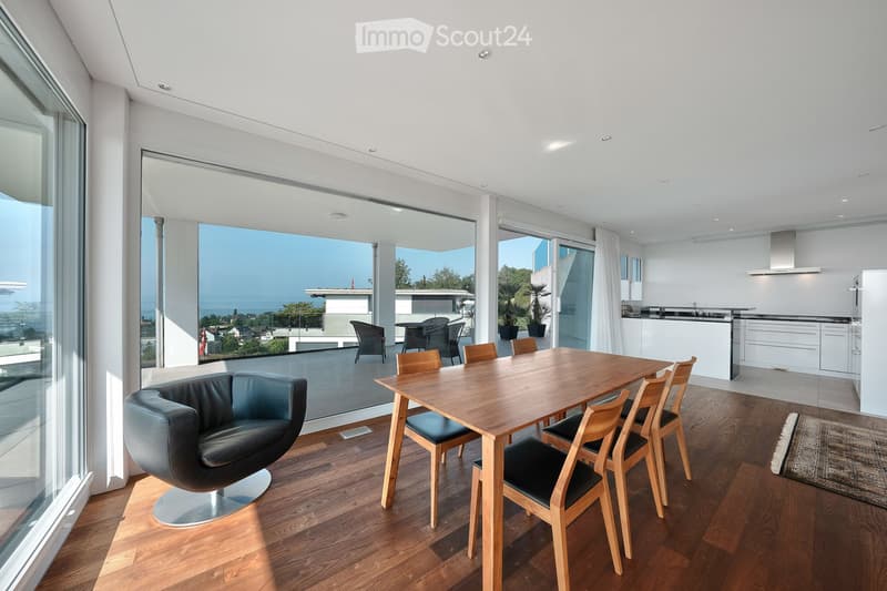 Modern dining area with a wooden dining table, black leather chair, and a view of the ocean through large glass windows. Adjacent kitchen area with white cabinets and a countertop.