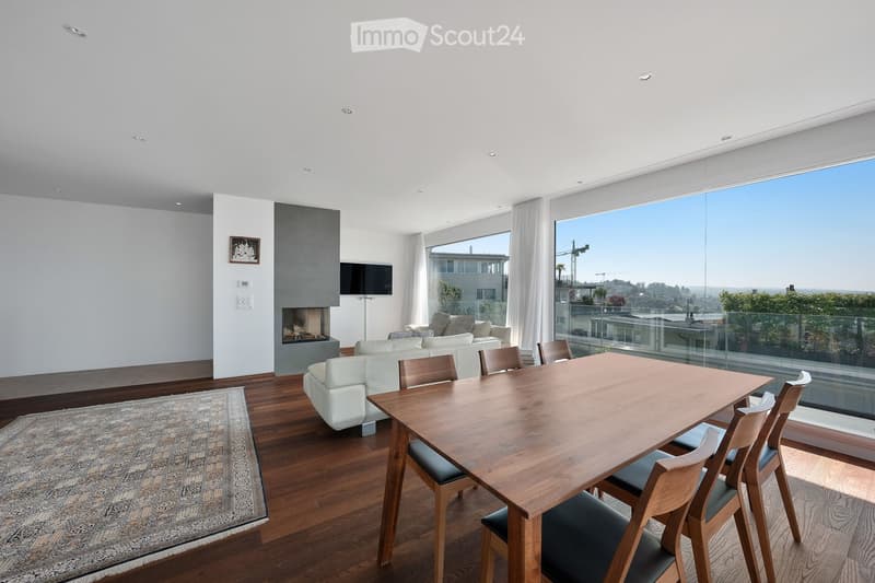 Modern living and dining area with white walls, wooden flooring, large windows offering a city view, a fireplace, a sofa, a dining table with chairs, and a television.
